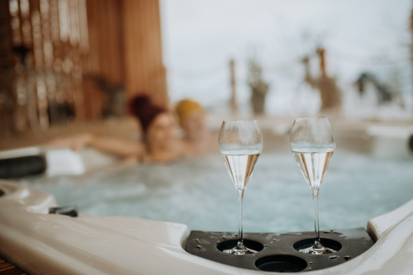 Senior couple in knitted cap enjoying together outdoor bathtub with glass of wine at their terrace during a cold winter day.