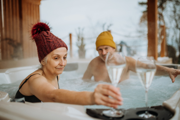 Senior couple in knitted cap enjoying together outdoor bathtub with glass of wine at their terrace during a cold winter day.