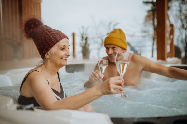 Senior couple in kintted cap enjoying together outdoor bathtub and clinking glasses at their terrace during a cold winter day.