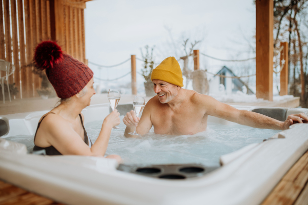 Senior couple in kintted cap enjoying together outdoor bathtub and clinking glasses at their terrace during a cold winter day.