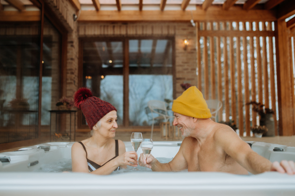 Senior couple in kintted cap enjoying together outdoor bathtub and clinking glasses at their terrace during a cold winter day.