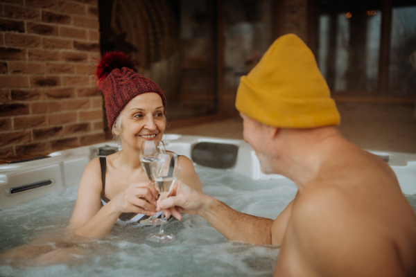 Senior couple in kintted cap enjoying together outdoor bathtub and clinking glasses at their terrace during a cold winter day.