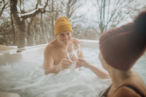 Senior couple in kintted cap enjoying together outdoor bathtub and clinking glasses at their terrace during a cold winter day.