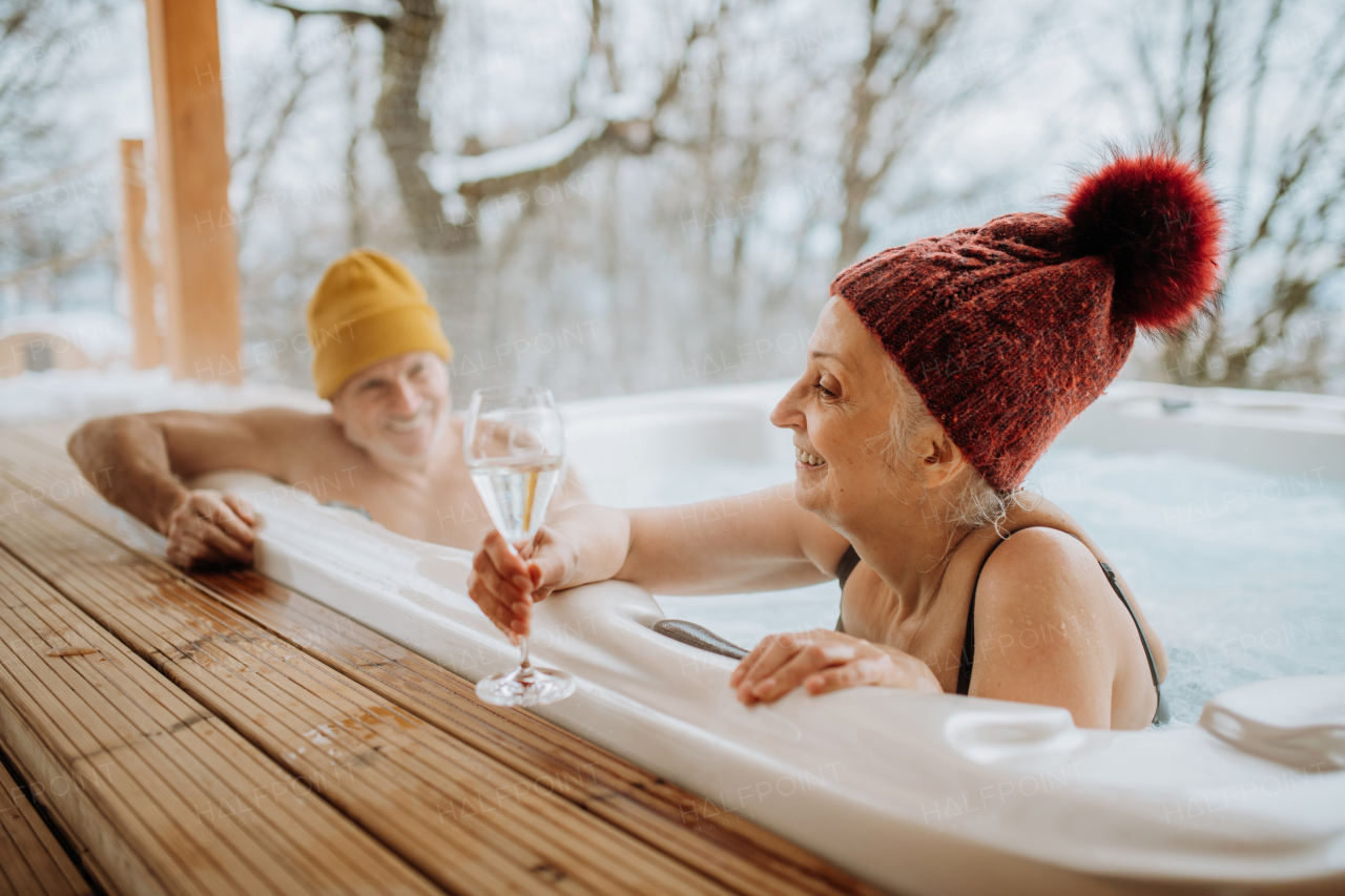 Senior couple in kintted cap enjoying together outdoor bathtub and clinking glasses at their terrace during a cold winter day.