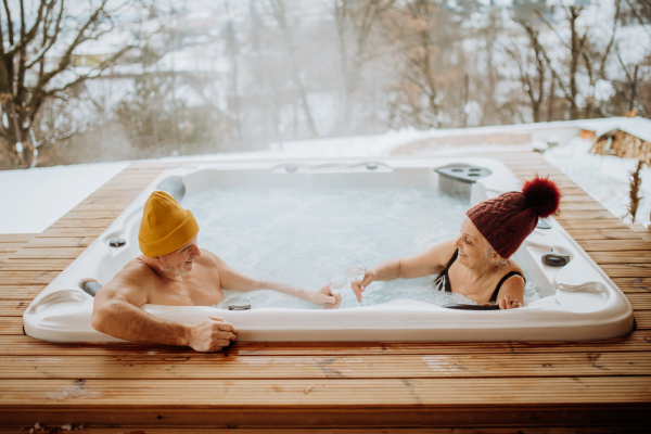 Senior couple in kintted cap enjoying together outdoor bathtub and clinking glasses at their terrace during a cold winter day.