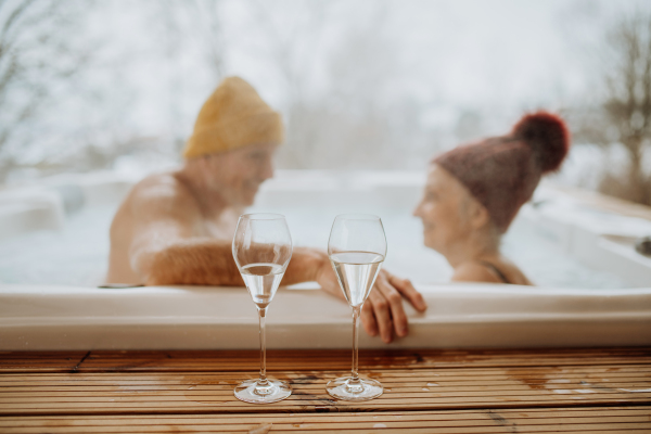 Senior couple in knitted cap enjoying together outdoor bathtub with glass of wine at their terrace during a cold winter day.