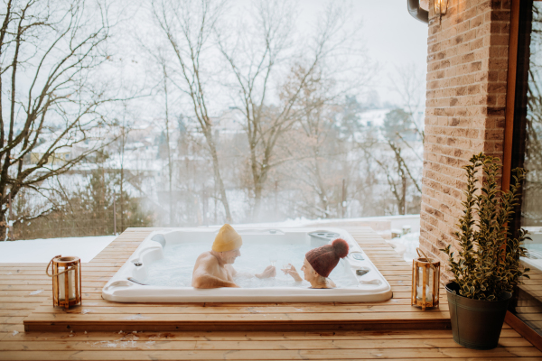 Senior couple in knitted cap enjoying together outdoor bathtub and view at their terrace during a cold winter day.