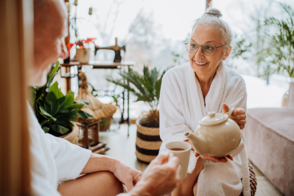 Senior couple in bathrobes enjoying time together in their living room, drinking a hot tea, calm and hygge atmosphere.