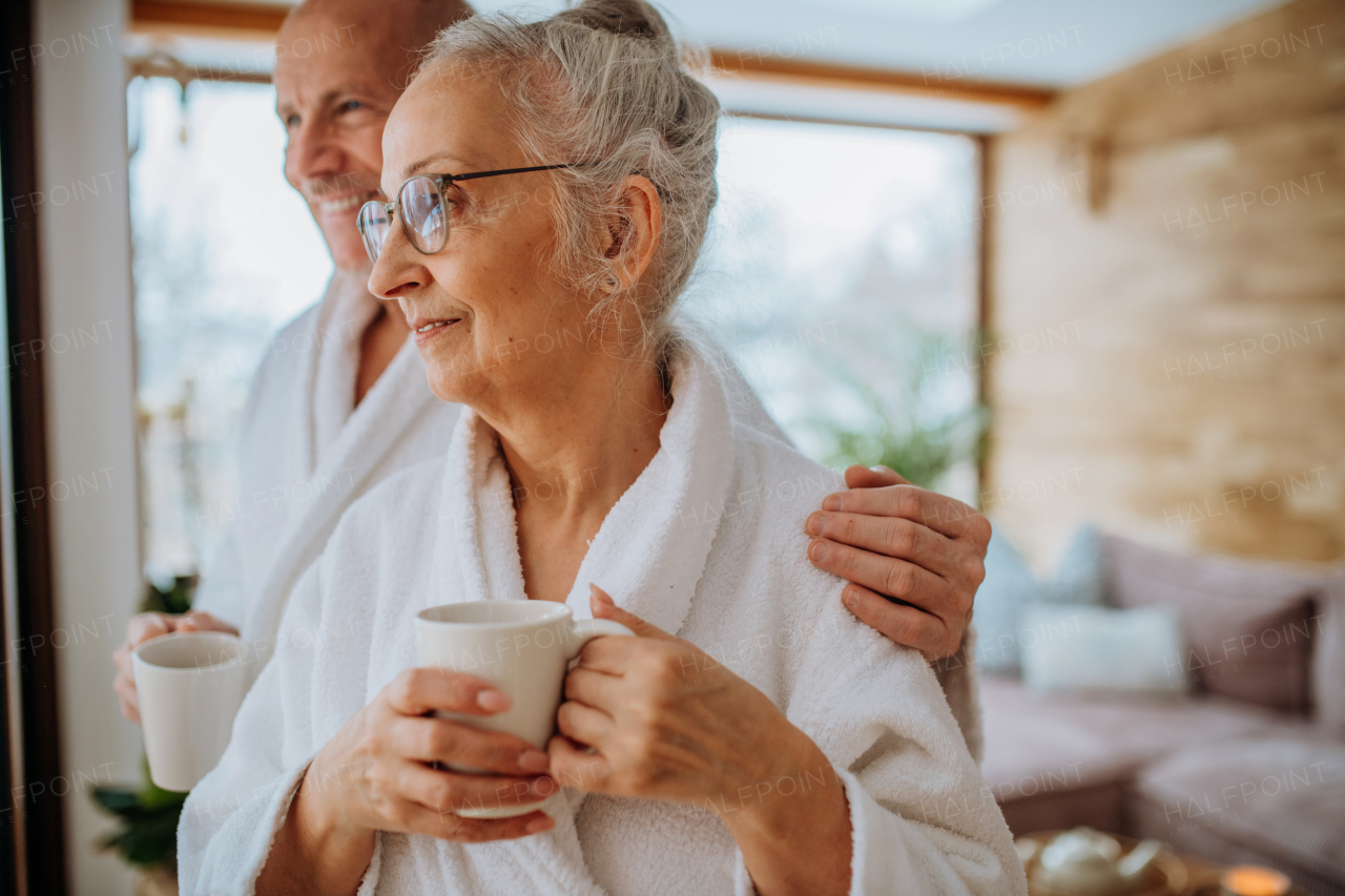 Senior couple in bathrobes enjoying time together in their living room, drinking a hot tea, calm and hygge atmosphere.