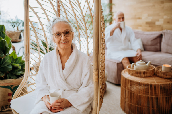 Senior woman sitting on indoor swing and looking at camera, her husband enjoying cup of tea in the background.