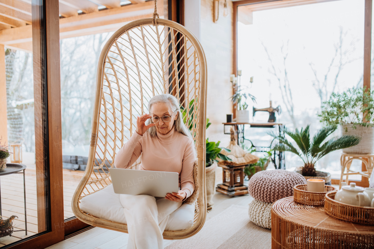 Senior woman sitting in indoor swing and working at a laptop. Concept of home office and active senior lifestyle.