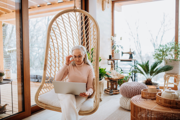 Senior woman sitting in indoor swing and working at a laptop. Concept of home office and active senior lifestyle.