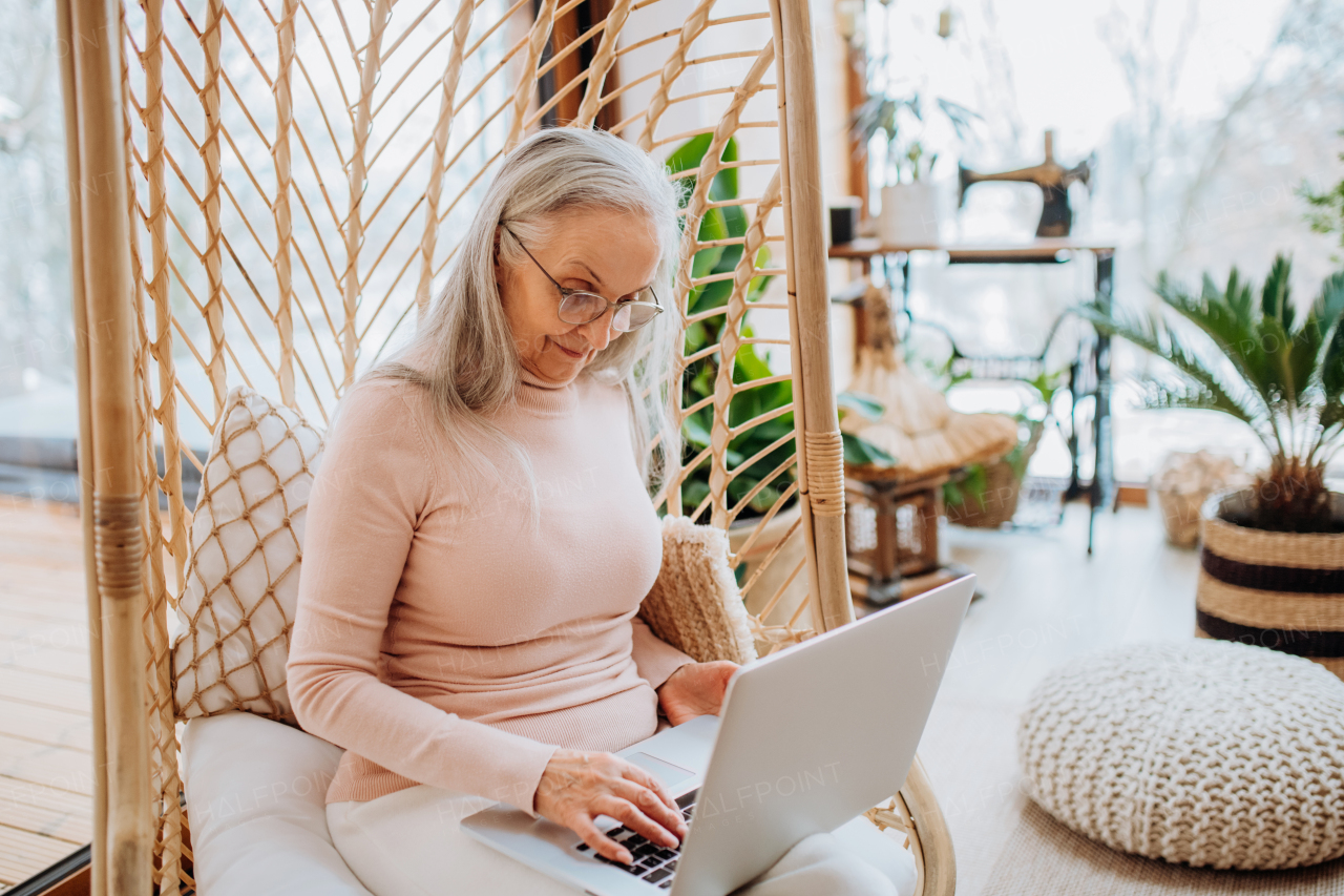 Senior woman sitting in indoor swing and working at a laptop. Concept of home office and active senior lifestyle.