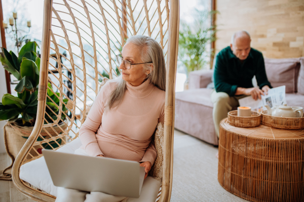 Senior woman sitting in outdoor swing and working at laptop, her husband resting at a sofa. Concept of home office and active senior lifestyle.