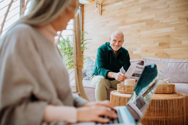 Senior woman sitting in indoor swing and working at laptop, her husband resting at a sofa. Concept of home office and active senior lifestyle.