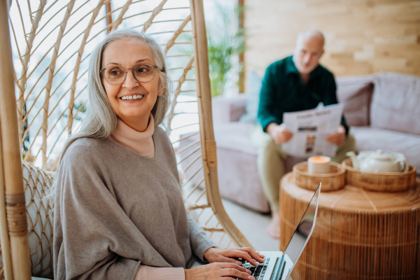 Senior woman sitting in outdoor swing and working at laptop, her husband resting at a sofa. Concept of home office and active senior lifestyle.