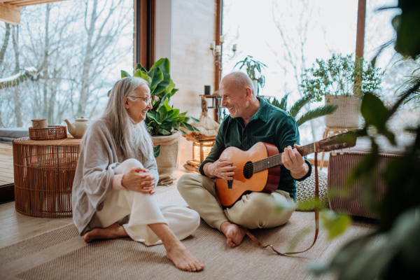 Senior man playing on guitair his wife, sitting in a cozy living room and enjoying autumn day.