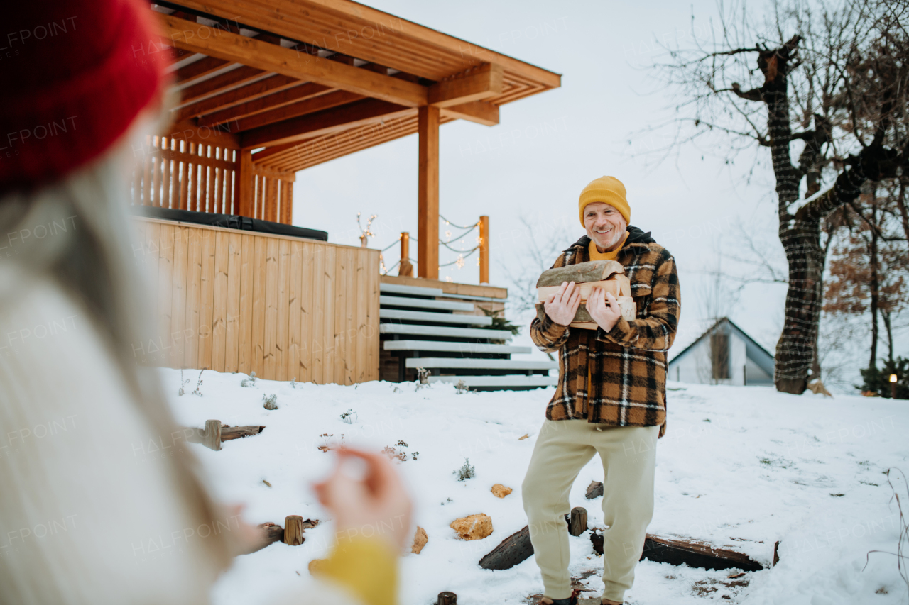 Senior man carrying wooden logs and preparing outdoor fire, during a winter day.