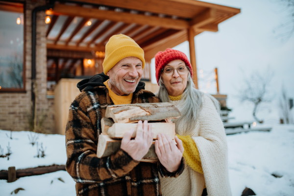 Senior couple holding wooden logs in front of their house, during a winter day.