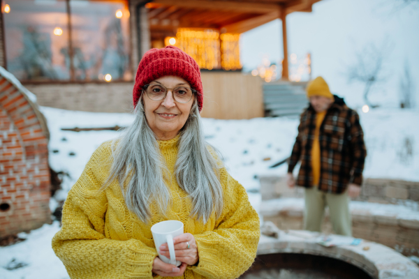 Happy senior woman with cup of hot tea standing in front of house during winter, her husband prepairing fire.