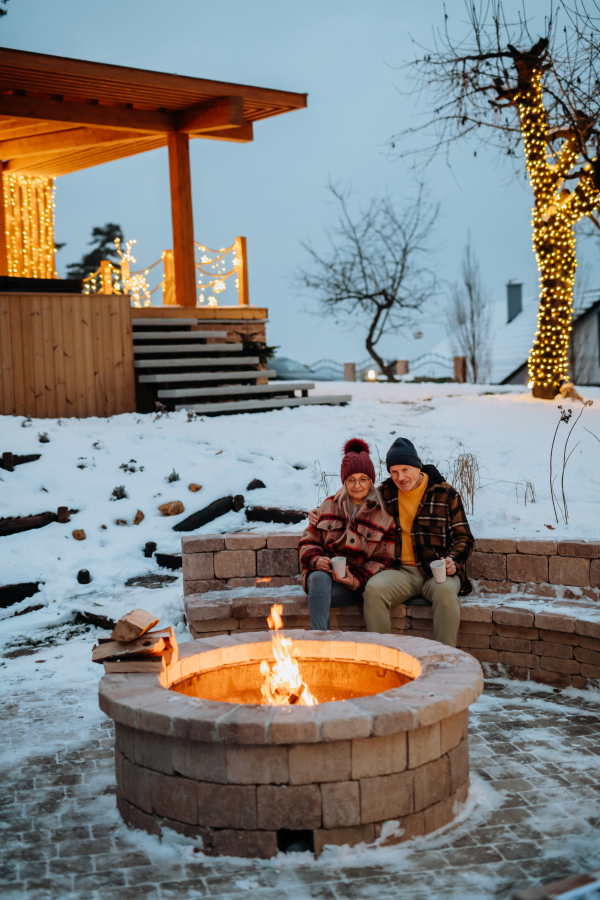 Senior couple sitting and heating together at outdoor fireplace during winter evening.