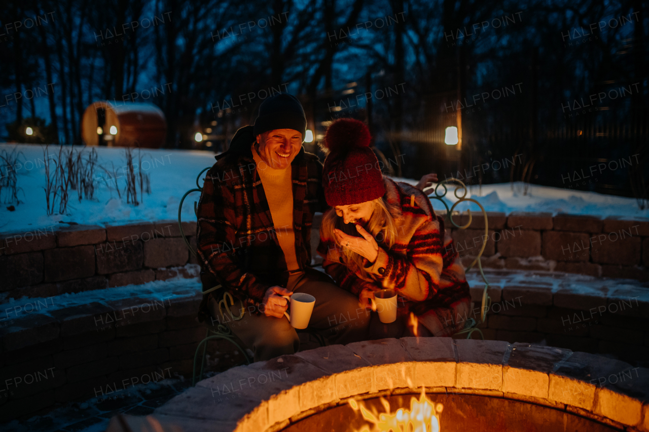 Senior couple sitting and heating together at outdoor fireplace during winter evening.