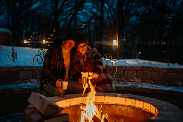 Senior couple sitting and heating together at outdoor fireplace during winter evening.