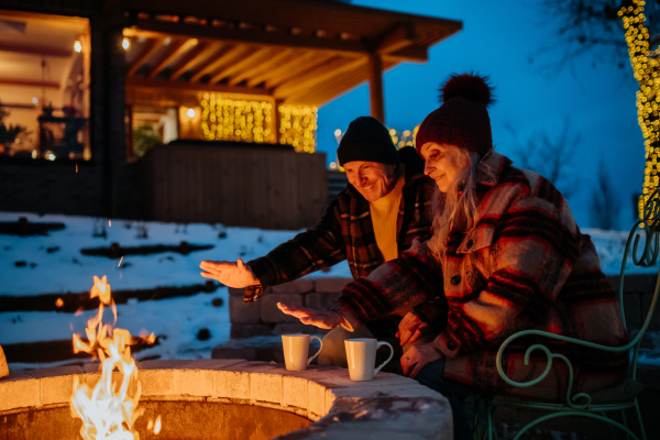 Senior couple sitting and heating together at outdoor fireplace during winter evening.