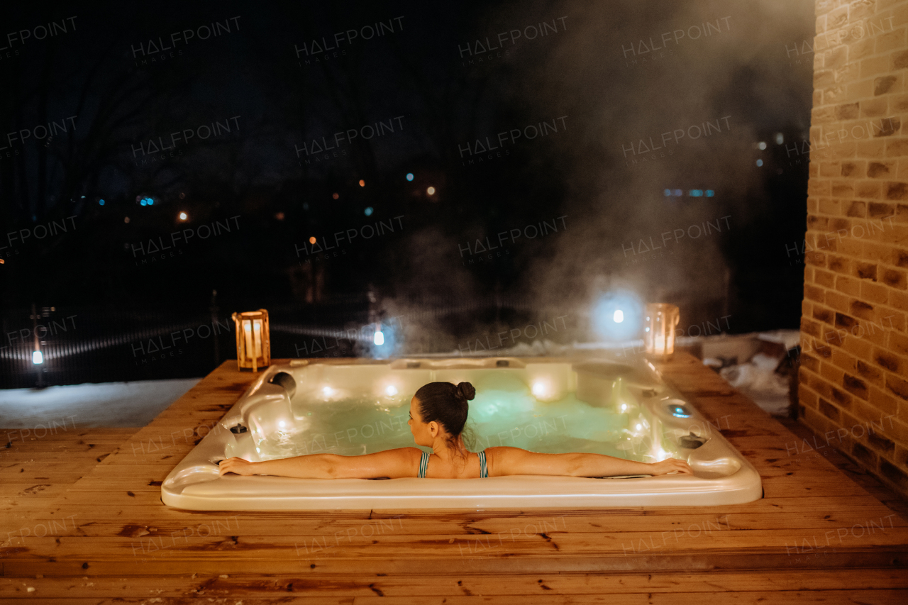 Rear view of young woman enjoying outdoor bathtub in her terrace during a cold winter evening.