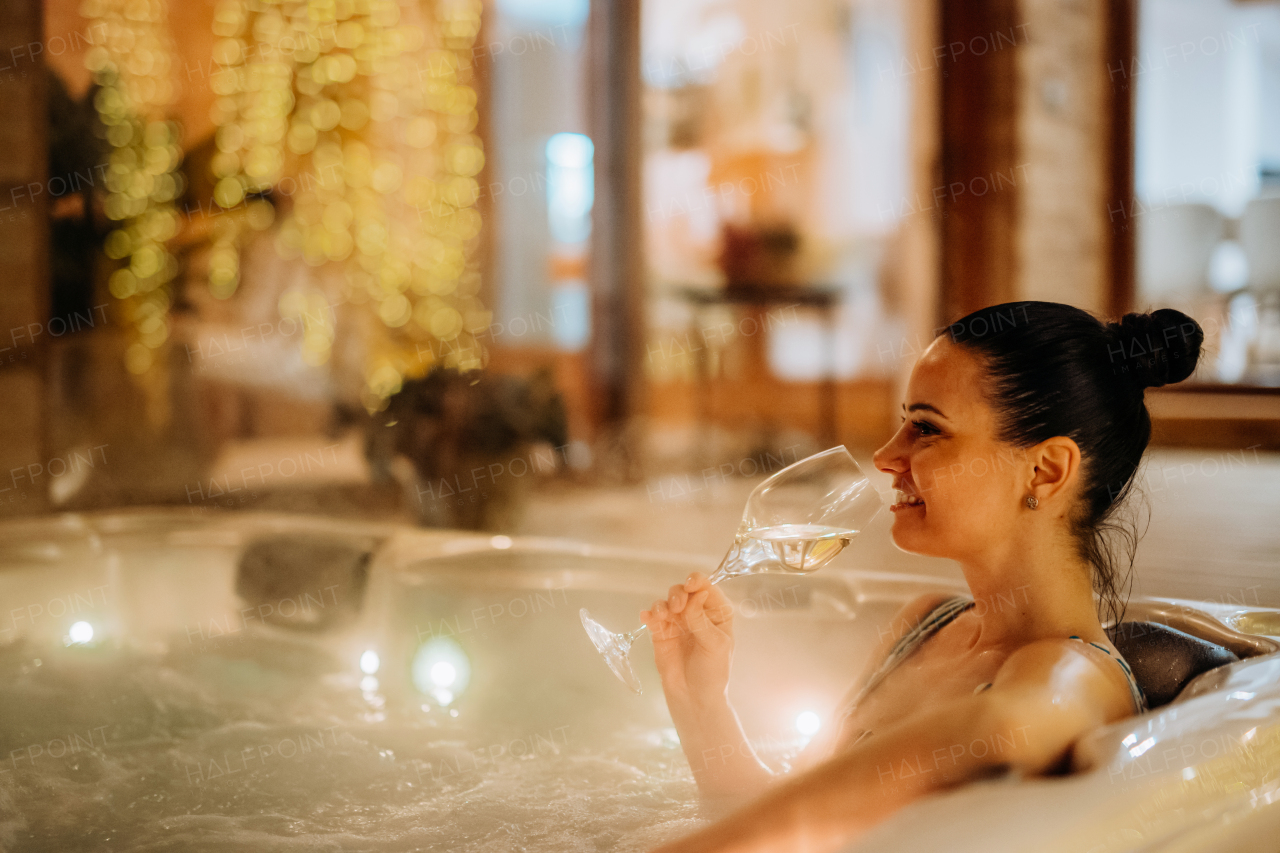 Young woman enjoying outdoor bathtub with glass of wine at her terrace during a cold winter evening.