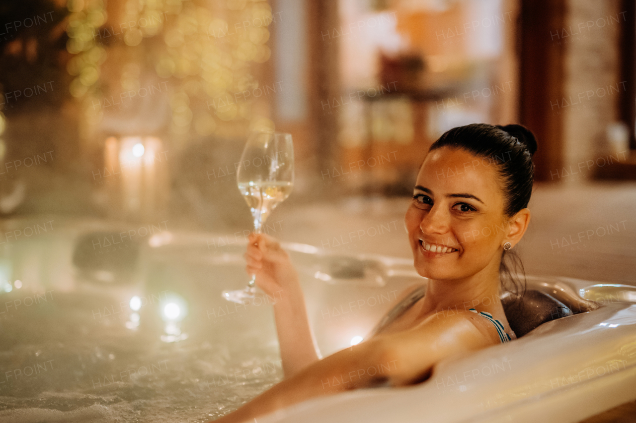 Young woman enjoying outdoor bathtub with glass of wine at her terrace during a cold winter evening.