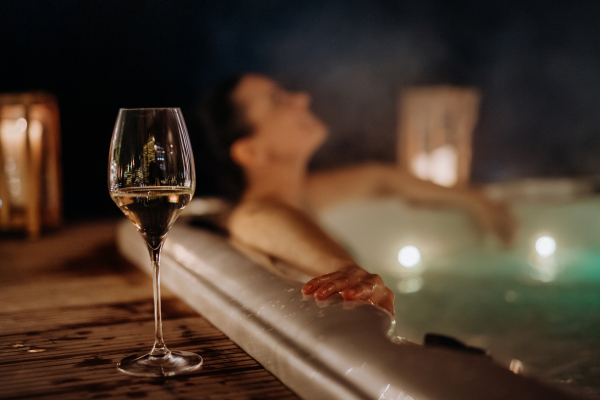 Young woman enjoying outdoor bathtub with glass of wine at her terrace during a cold winter evening.