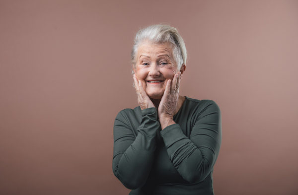 Studio portrait of smiling senior woman with short gray hair, holding her face. Isolated on a brown pink background. Copy space.