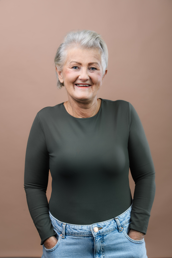 Studio portrait of smiling senior woman with hands in jeans pocket. short gray hair, green top. Isolated on a brown pink background. Copy space.