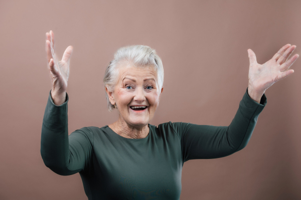 Studio portrait of smiling senior woman with raised arms. short gray hair, green top. Isolated on a brown pink background. Copy space.