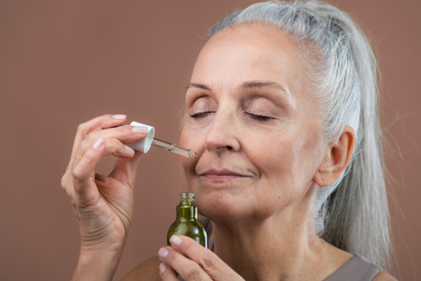 Studio portrait of beautiful senior woman holding skincare serum drops in bottle. Elderly woman taking care of aging skin. Concept of beauty and skincare.