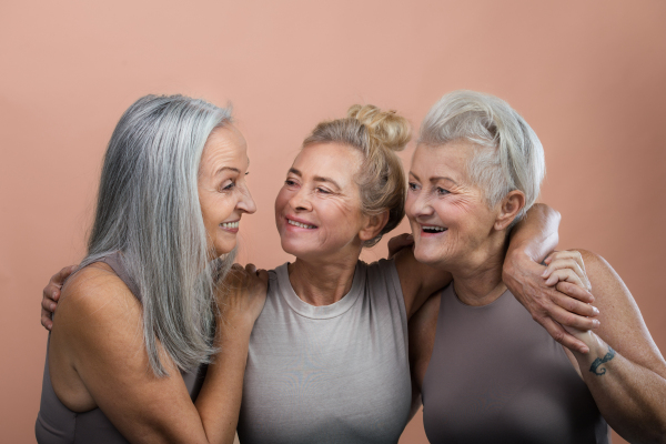 Studio portrait of three beautiful senior women with gray hairs. Elderly female friends holding each other and smiling. Isolated on a brown pink background. Copy space.