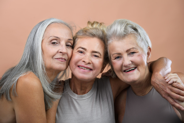 Studio portrait of three beautiful senior women with gray hairs. Elderly female friends embrace each other looking at camera. Isolated on a brown pink background. Copy space.