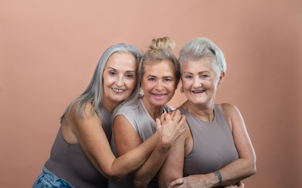 Studio portrait of three beautiful senior women with gray hairs. Elderly female friends holding each other looking at camera. Isolated on a brown pink background. Copy space.