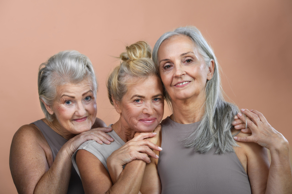 Studio portrait of three beautiful senior women with gray hairs. Elderly female friends holding each other looking at camera. Isolated on a brown pink background. Copy space.