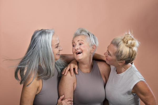 Studio portrait of three beautiful senior women with gray hairs. Elderly female friends holding each other, laughing. Isolated on a brown pink background. Copy space.