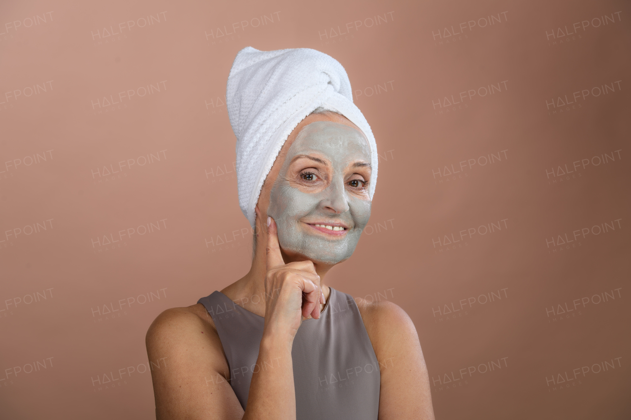 Portrait of beautiful senior woman with a face-mask and hair turban towel. Selfcare and skin routine concept. Isolated on a brown pink background. Copy space.