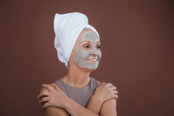 Portrait of beautiful senior woman with a face-mask and hair turban towel. Selfcare and skin routine concept. Isolated on a brown pink background. Copy space.