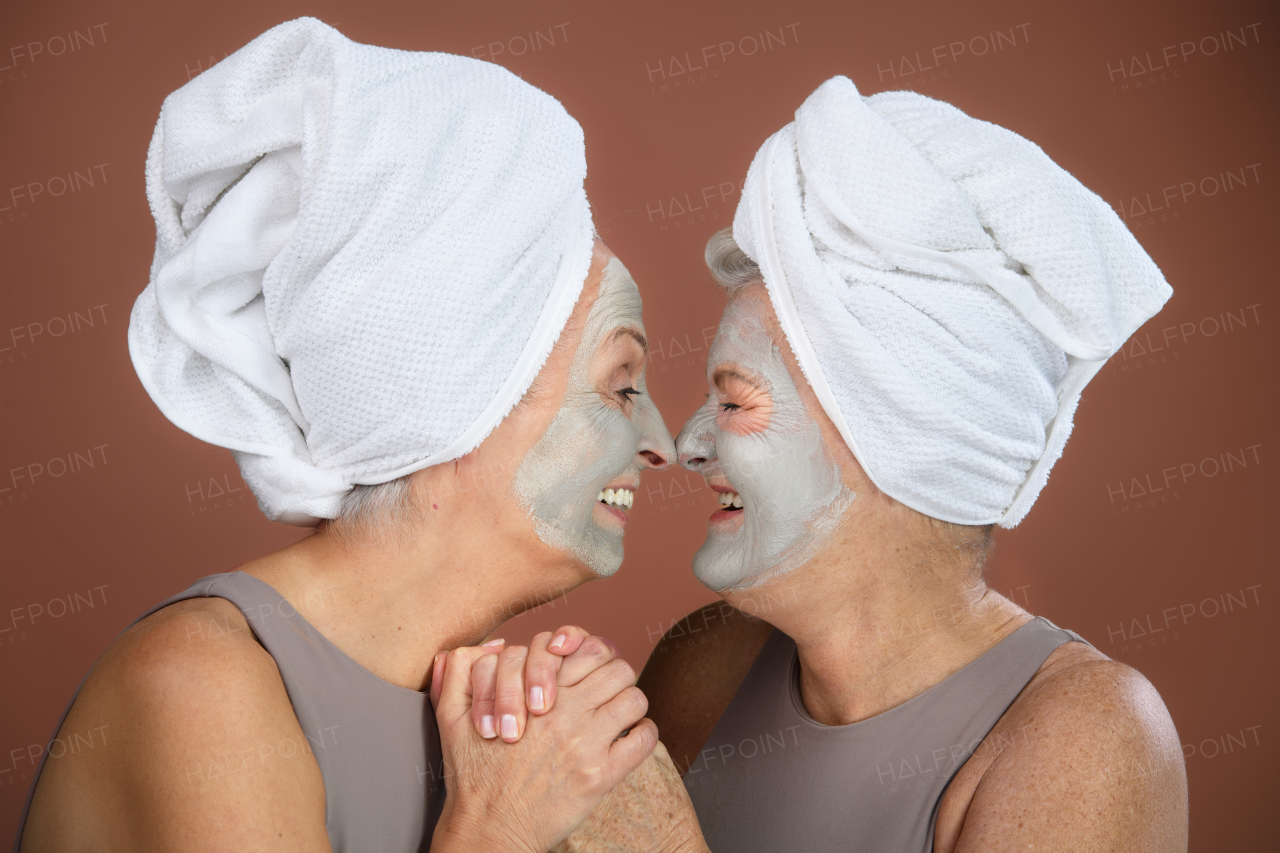 Portrait of happy senior women with a face-mask and hair turban towel, looking at each other, holding hands. Selfcare and skin routine concept. Isolated on a brown pink background. Copy space.