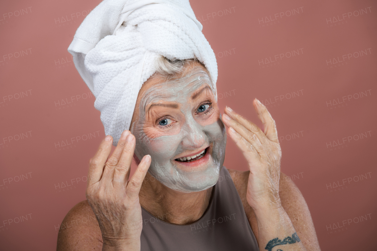 Portrait of smiling senior woman with a face-mask and hair turban towel. Selfcare and skin routine concept. Isolated on a brown pink background. Copy space.