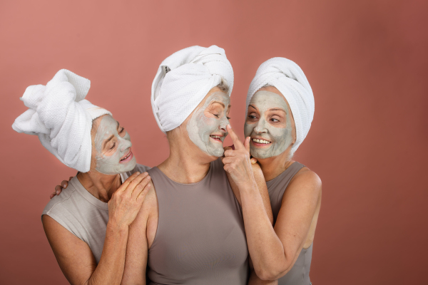 Portrait of three happy senior women with a face-mask and hair turban towel, taking care of aging skin. Selfcare and skin routine concept. Isolated on a brown pink background. Copy space.