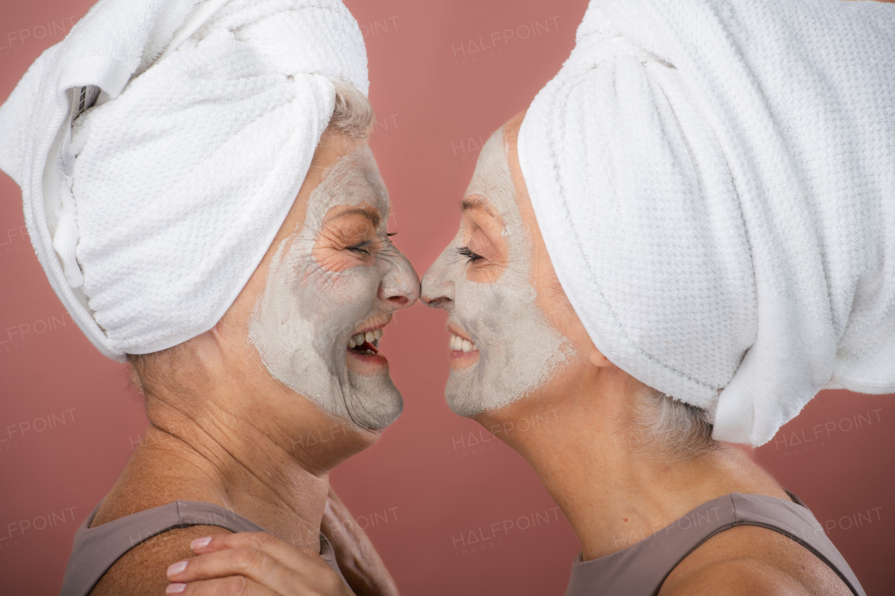 Portrait of happy senior women with a face-mask and hair turban towel, looking at each other. Selfcare and skin routine concept. Isolated on a brown pink background. Copy space.