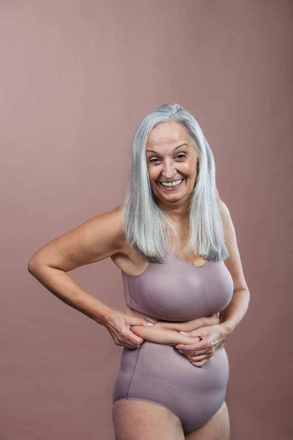 Portrait of senior woman in a lingerie, studio shoot. Beautiful body shape of aging woman, signs of age on woman body. Isolated on a brown pink background. Copy space.