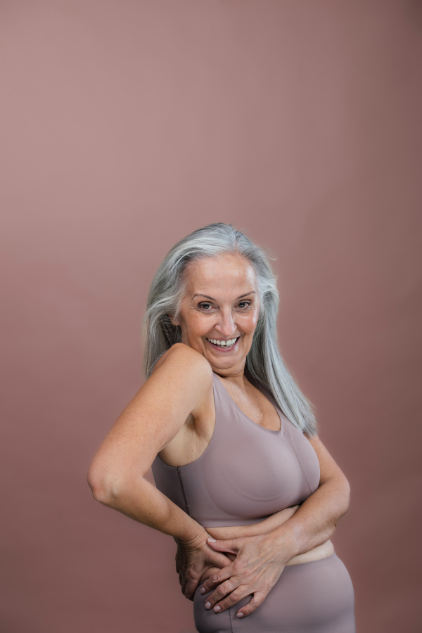 Portrait of senior woman in a lingerie, studio shoot. Beautiful body shape of aging woman. Isolated on a brown pink background. Copy space.