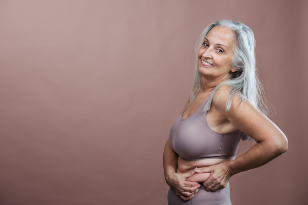 Portrait of senior woman in a lingerie, studio shoot. Beautiful body shape of aging woman, signs of age on woman body. Isolated on a brown pink background. Copy space.